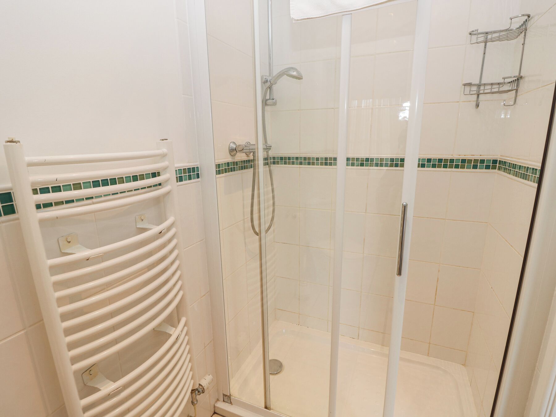 Una ducha con cerramiento de vidrio, pared con azulejos blancos, toallero blanco y detalle decorativo con azulejos verdes.