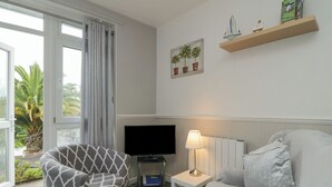 A living room with a sofa, a chair, a TV, a lamp, and a window with curtains. - Flat 2, Big Tree Holiday Flats (Paignton)