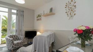 A hotel room with a sofa, a TV, a dining table with plates and glasses, a vase with flowers, and a window with curtains. - Flat 2, Big Tree Holiday Flats (Paignton)