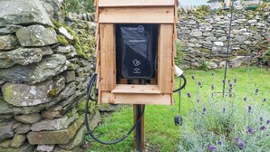 Une structure en bois, dotée d’un panneau solaire noir et d’un port de chargement, entourée de murs en pierre et de verdure.