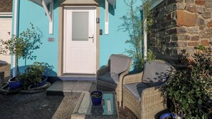 A building with a white door, a blue exterior, and a stone wall. There are wicker chairs and potted plants outside. - The Hollybush (Kilgetty)