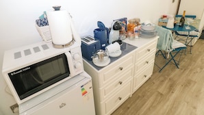 A kitchenette with a microwave, toaster, and coffee maker. There is a table with a bottle of wine, glasses, and a box of cereal. A chair and a small table are also visible. - The Hollybush (Kilgetty)