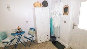 A room with a white wardrobe, a small table with a bottle and glasses, a blue folding chair, and a door with a mat. - The Hollybush (Kilgetty)
