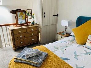 A bedroom with a bed, a dresser, a nightstand, a lamp, and a window with blinds. - Cockle Bay Cottage (Morecambe)