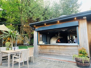 Un four à pizza extérieur, avec un chef à l’intérieur, entouré de tables et de chaises en bois, et des guirlandes lumineuses suspendues au-dessus.