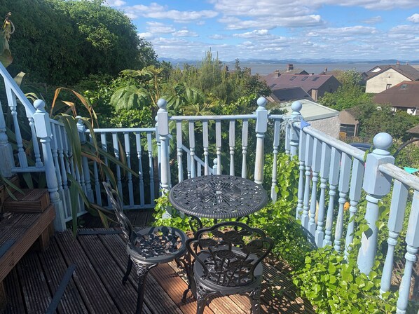 A wooden deck with a metal table and chairs, surrounded by a white railing with a view of houses and a body of water in the background. - Cockle Bay Cottage (Morecambe)