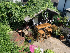 A garden with a wooden pergola, a small table, and a chair. There are potted plants and a sign with flags. - Cockle Bay Cottage (Morecambe)