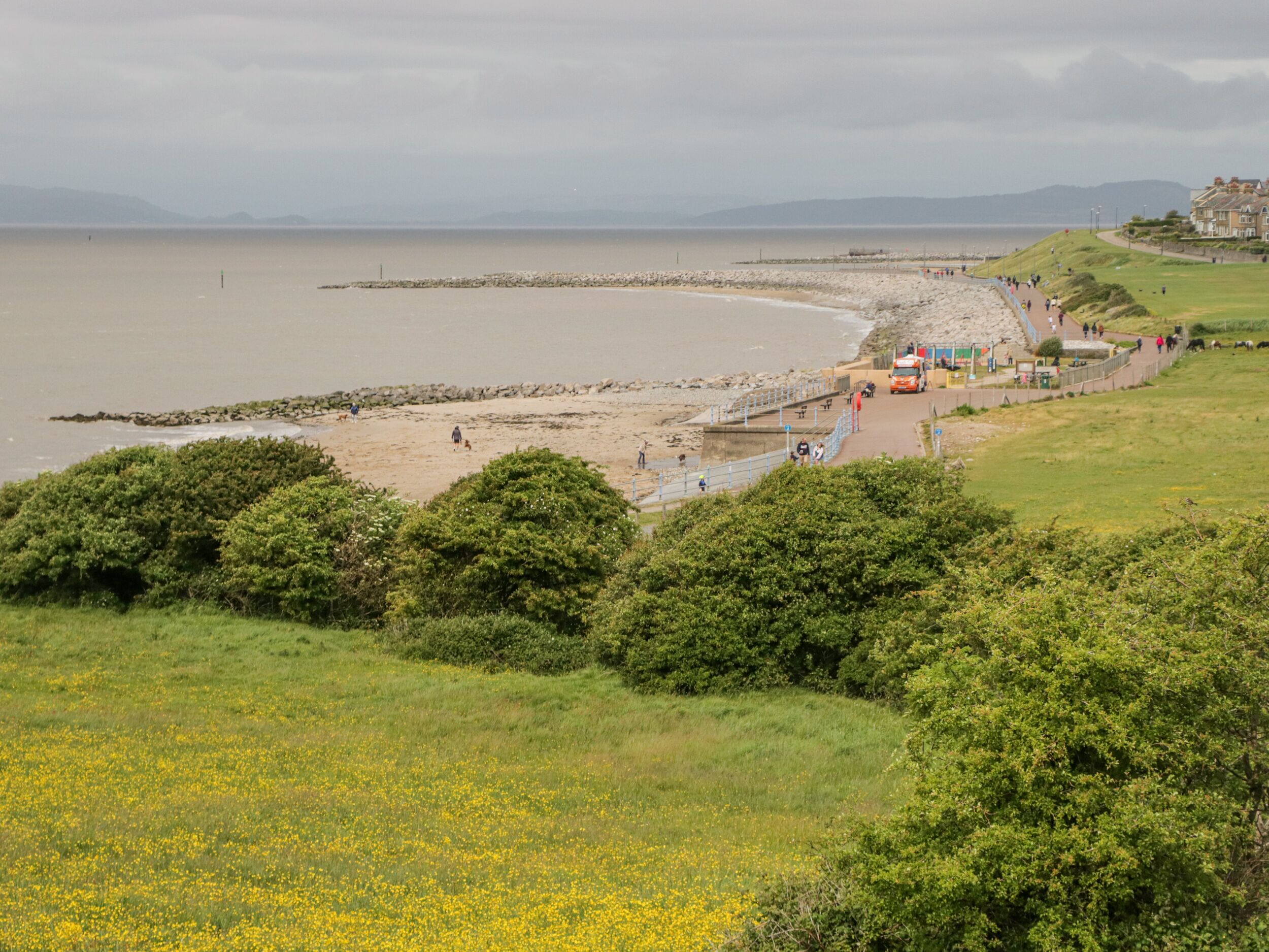 Ein Küstenstrand mit einer Steinmole, Grünflächen und einigen Gebäuden im Hintergrund.