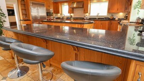 A modern kitchen with wooden cabinets, a large island with bar stools, stainless steel appliances, and a window providing natural light. - The Wing (Wrexham)