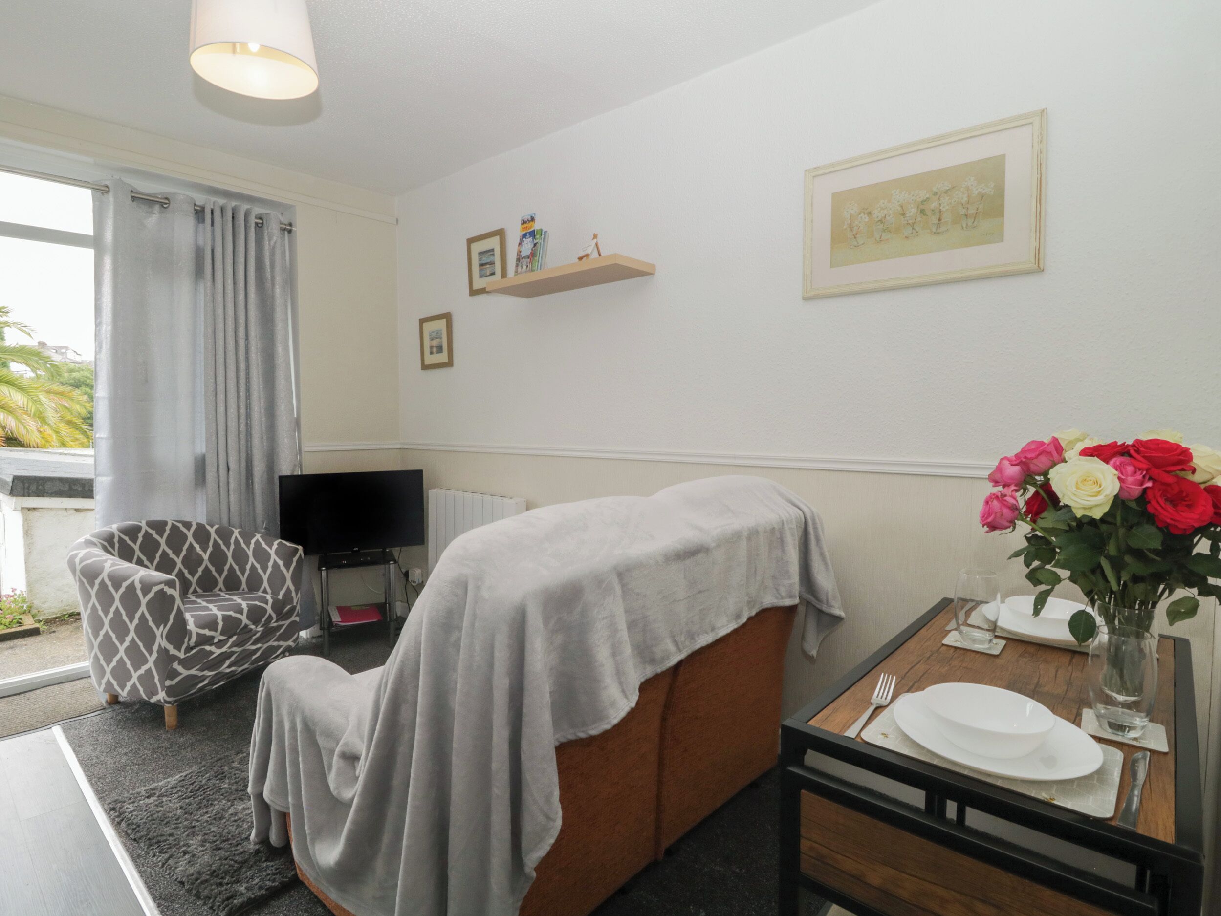 A room with a sofa covered with a grey blanket, a TV on a stand, a small table with a vase of flowers, and a window with curtains.