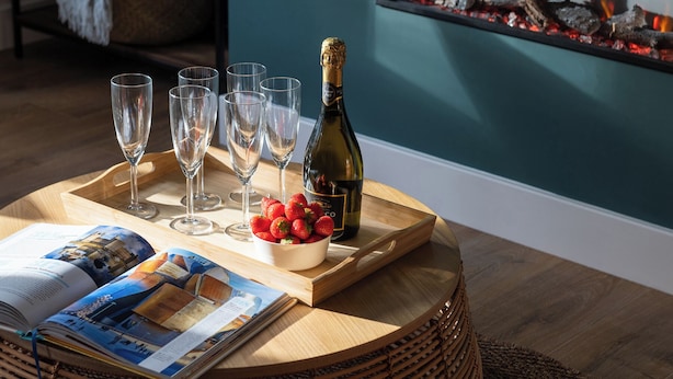 Een houten dienblad met een champagnefles, een schaal vol aardbeien en verschillende glazen op een ronde tafel.