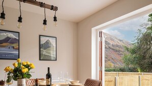 Een eetruimte met een houten tafel, bruine lederen stoelen, een fles wijn en een vaas met gele bloemen. Twee ingelijste foto's hangen aan de muur en er is uitzicht op een bergachtig landschap door een groot raam.