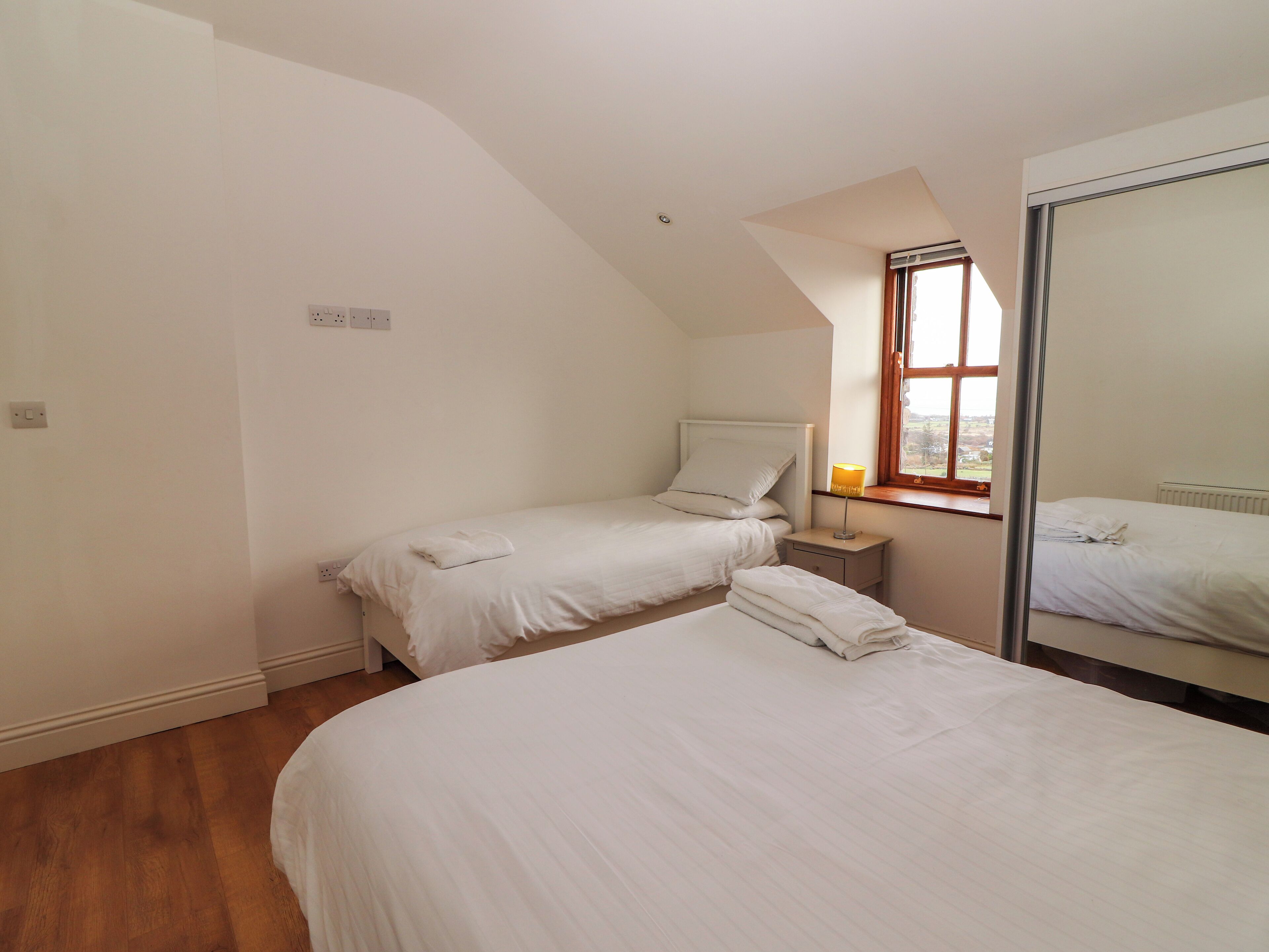 Een slaapkamer met twee eenpersoonsbedden, een nachtkastje met lamp, een raam met uitzicht en een spiegel aan de muur.