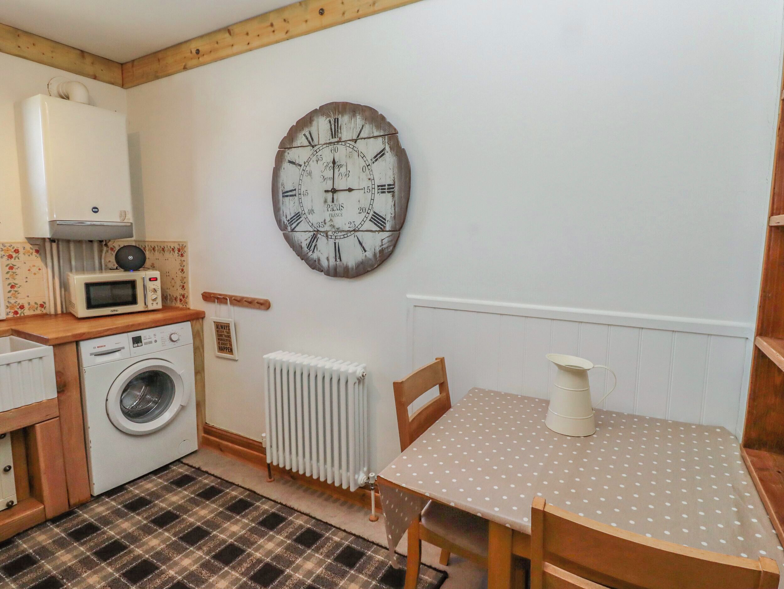 A compact kitchen with a washing machine, a microwave, a radiator, and a dining table with a polka dot tablecloth.