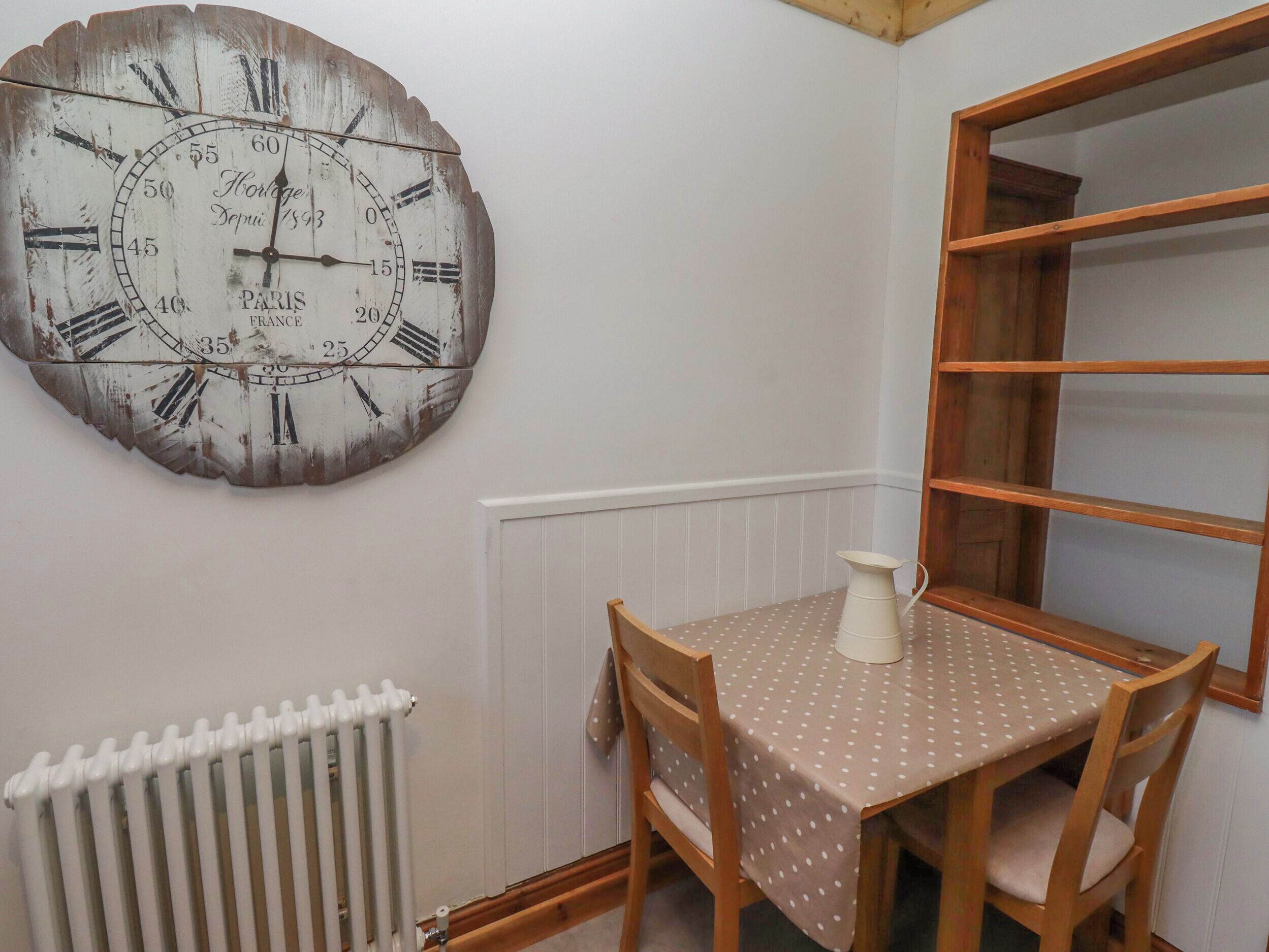 A rustic wooden wall clock with Roman numerals and a vintage design, a wooden table with a polka dot tablecloth, and a white radiator.