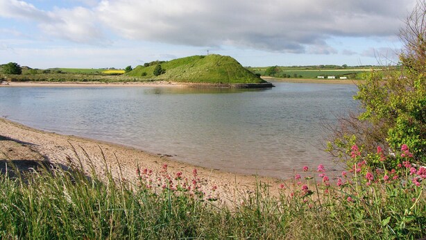 A river with a mound in the center, surrounded by greenery and a cloudy sky. - Seafield Granary (Alnwick)
