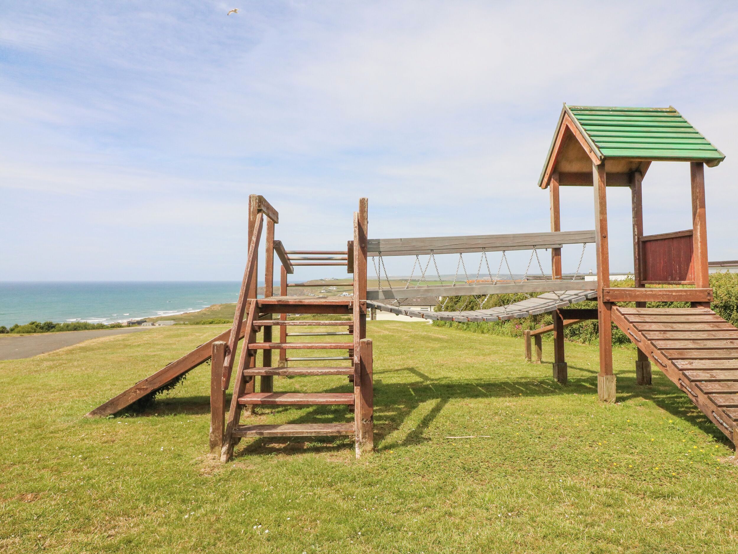 A wooden swing set with a green roof, a wooden ladder, and a grassy area with a view of the ocean.