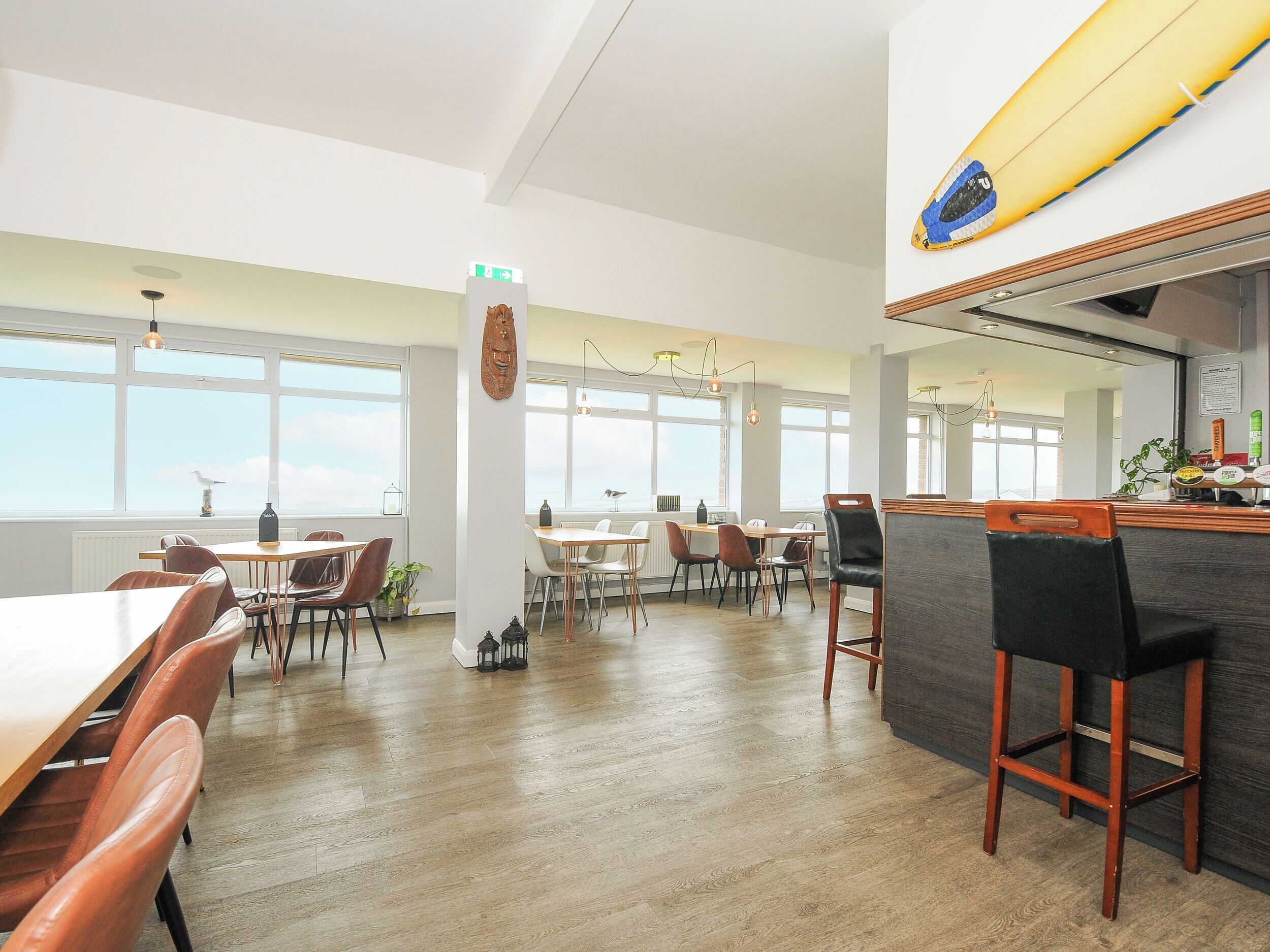 A spacious dining area with wooden flooring, multiple tables and chairs, and large windows offering a view of the sky.