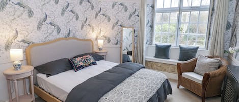 A bedroom with a large bed, a nightstand, a chair, a window with curtains, and a stone wall.