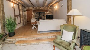 A cozy living room with a stone fireplace, a sofa, a green armchair, and a television. - Lower Pack Horse Cottage (Cheltenham)