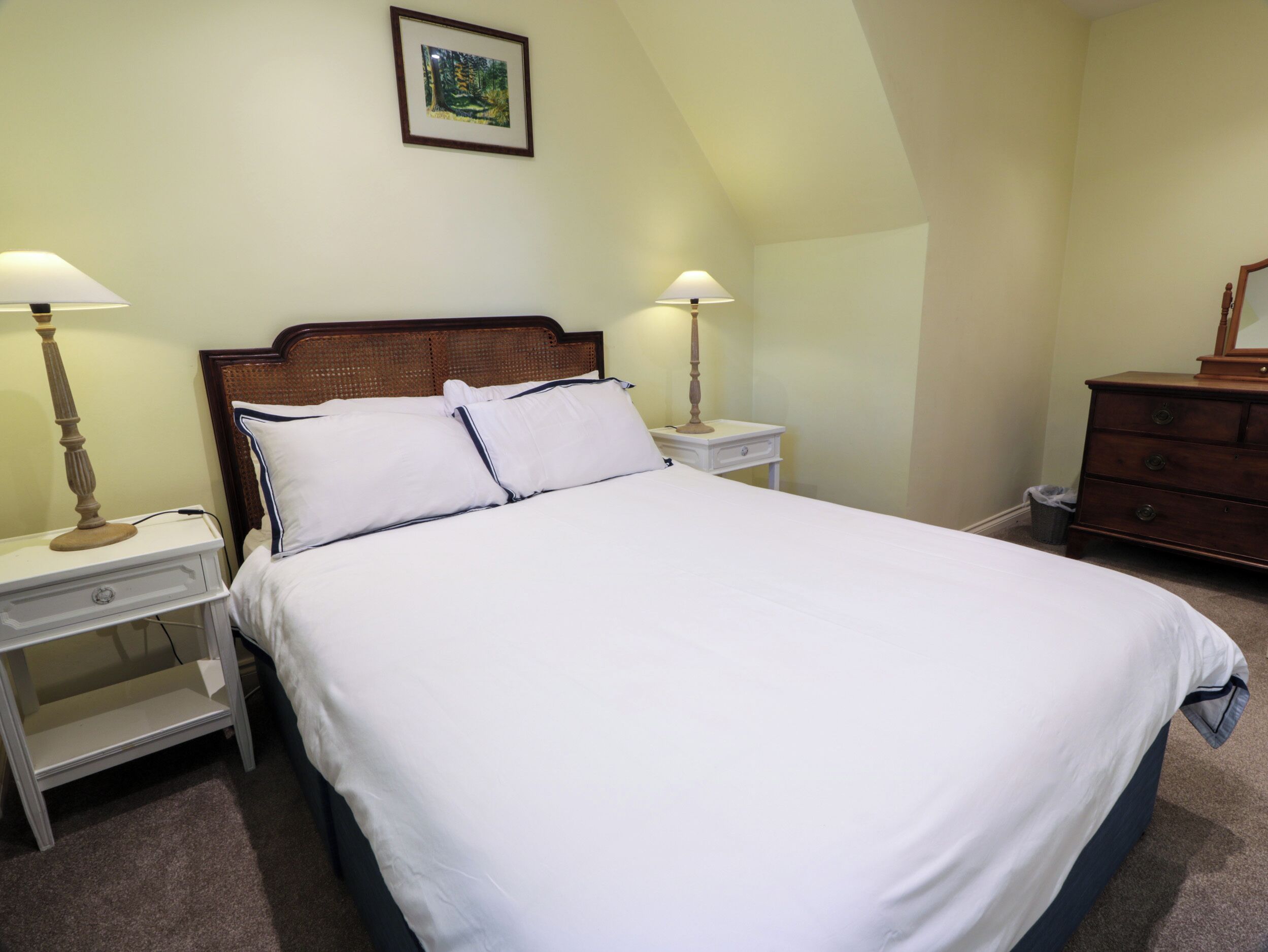 A neatly made bed with white linens and navy blue trim, flanked by two bedside tables with lamps, in a room with a sloped ceiling and a framed picture on the wall.