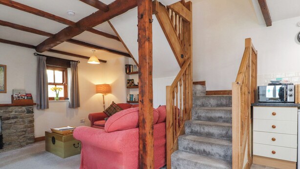 A cozy living room with a stone fireplace, a pink sofa, a wooden staircase, and a kitchen area with a toaster. - Mallards Nest (Skipton)