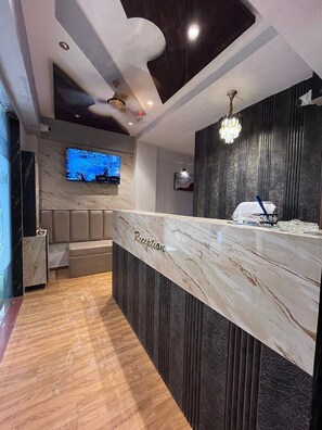 A reception area with a marble counter labeled "Reception," a television mounted on the wall, and a chandelier. - Hotel Privilon (Ahmedabad)