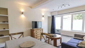 A living room with a dining table, chairs, a sofa, a TV on a wooden cabinet, and a large window with curtains. - West Morton Suite (Thornhill)