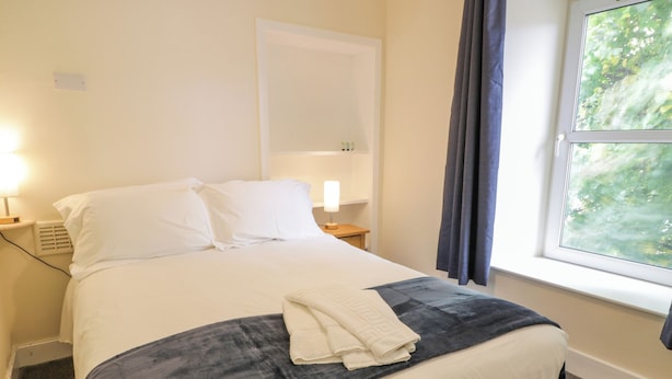 Una cama bien hecha con ropa de cama blanca y gris, una toalla blanca doblada y una ventana con cortinas.