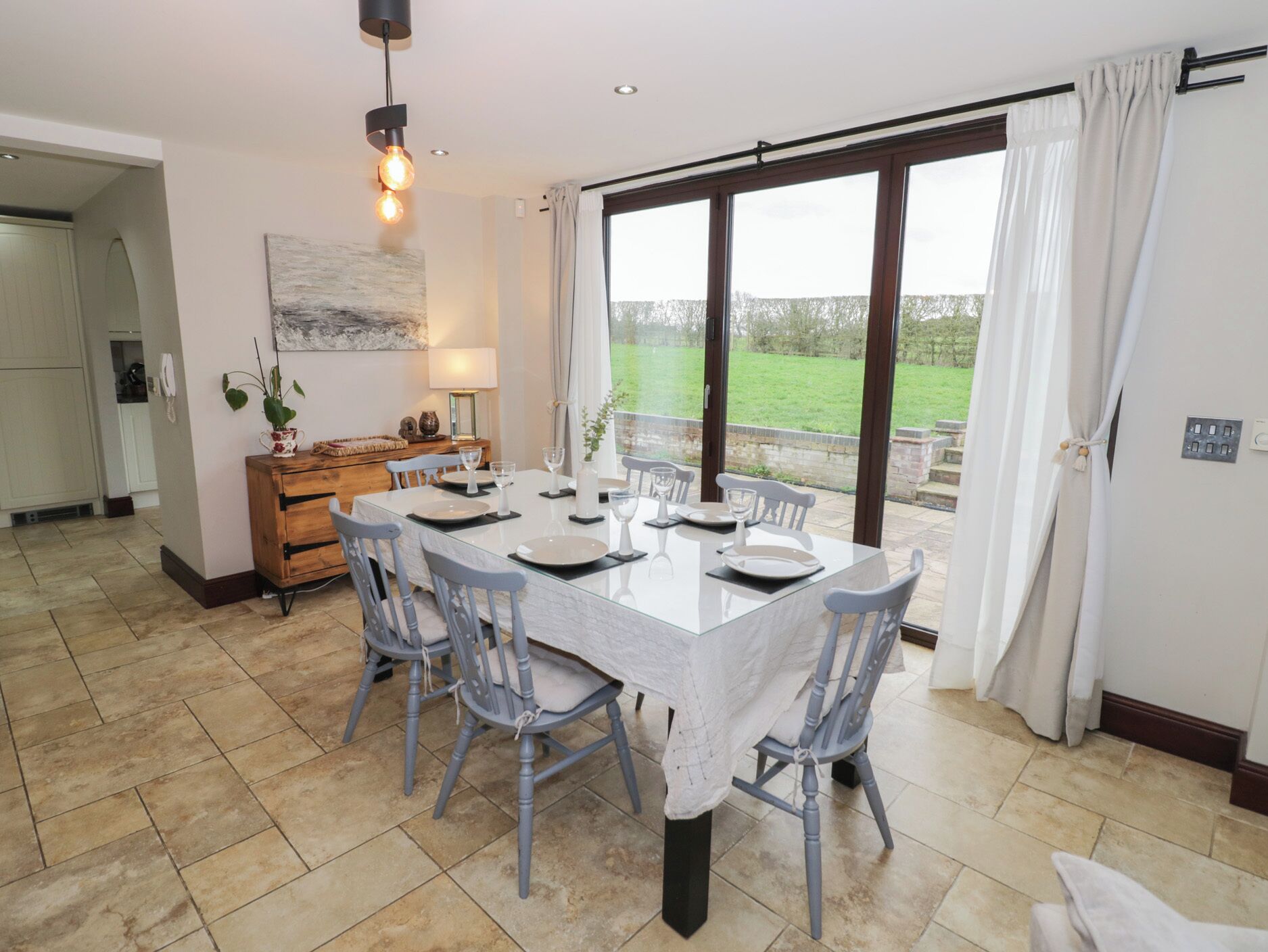 A dining area with a glass table, grey chairs, and a view of a garden through large windows.