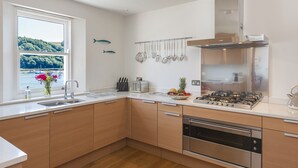Een moderne keuken met houten kastjes, roestvrijstalen apparatuur, een raam met uitzicht op zee en een fruitschaal op het aanrecht.