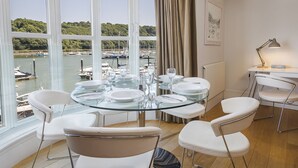 Een eetgedeelte met een glazen tafel, witte stoelen en uitzicht op een jachthaven met boten.