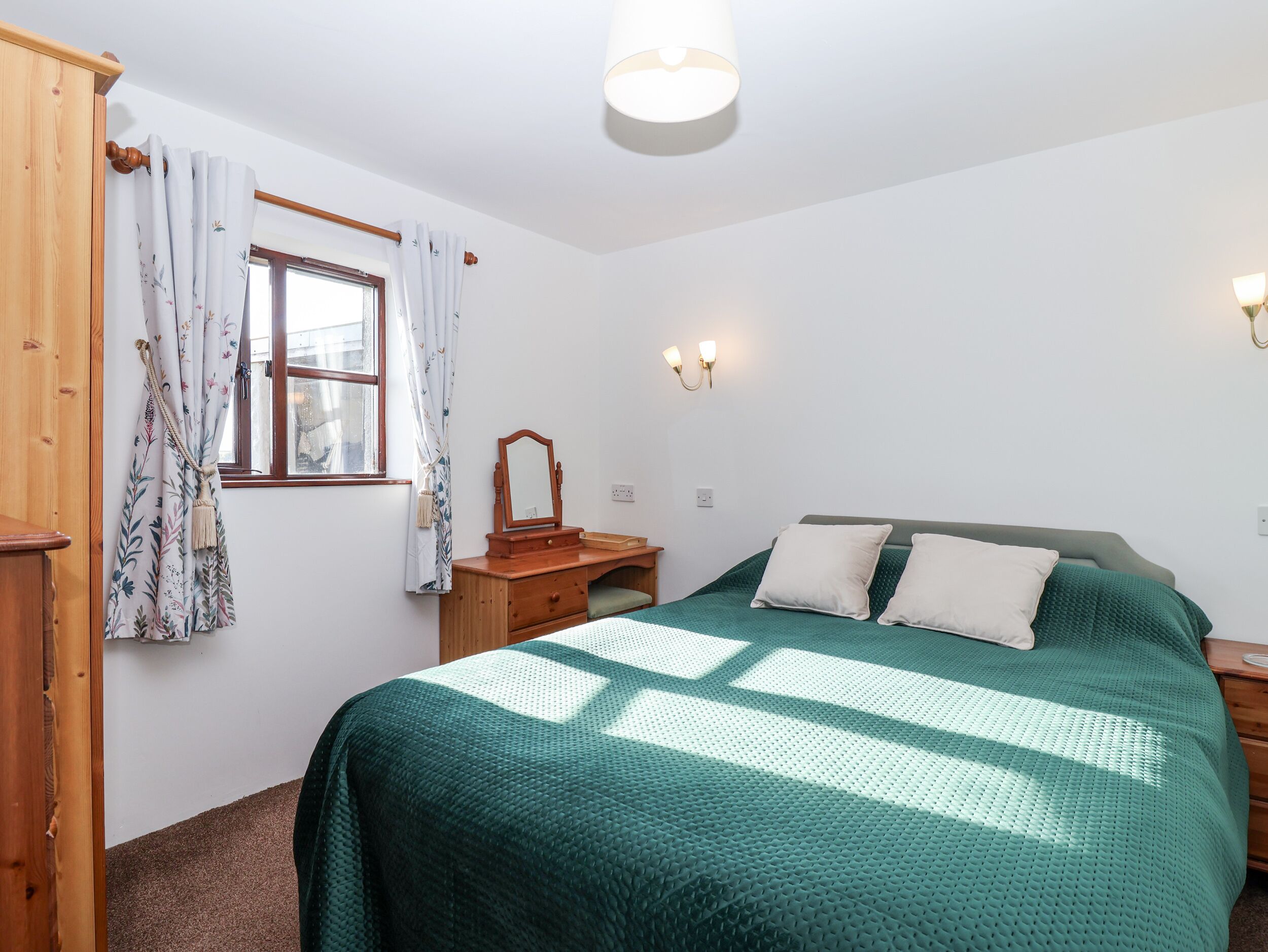 A bedroom with a bed, a wooden dresser, a window with curtains, and wall-mounted lights.