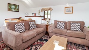 A living room with two brown sofas, a wooden coffee table, a patterned rug, a dining table with chairs, and a ceiling light. - The Dairy (Liskeard)