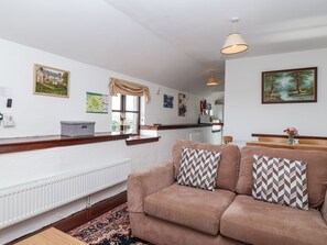 A cozy living room with a brown sofa, patterned cushions, a radiator, a window with a view, and a dining area in the background. - The Dairy (Liskeard)