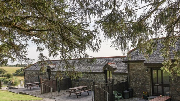 A stone building with a sloped roof, outdoor seating, and a view of a body of water. - The Dairy (Liskeard)