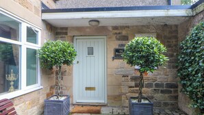 A stone building with a white door, two potted plants, and a bench. - The Old Coach House (Buxton)