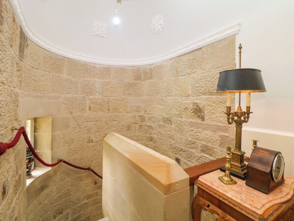 A stone-walled bathroom with a wooden bench, a red rope, a wall-mounted light fixture, and a table with a clock and a lamp.