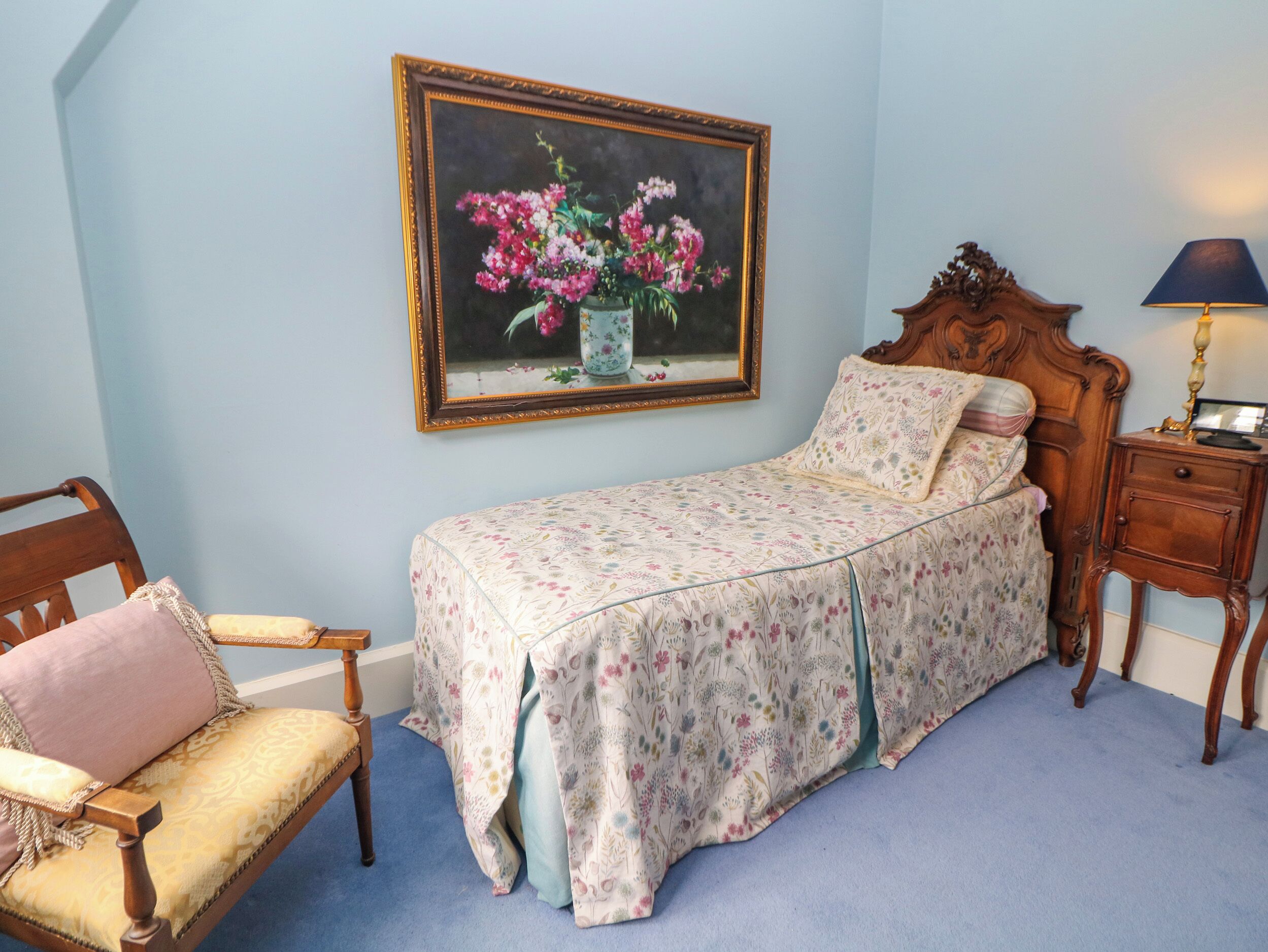 Un dormitorio con cama, una silla, una mesita de noche, una lámpara y un cuadro con flores en la pared.