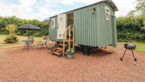 A small green shed with a door open, a set of stairs leading up to it, a table with chairs, and a barbecue grill.