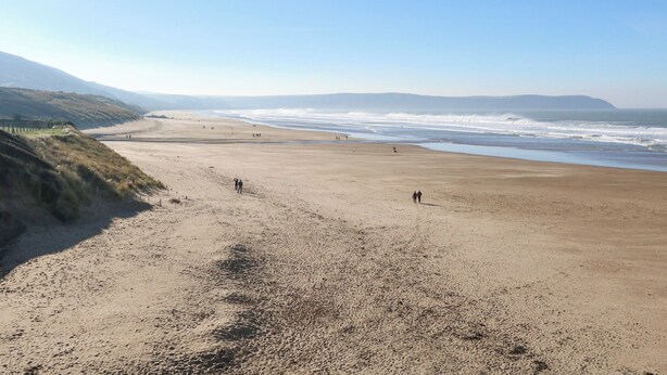 A wide sandy beach with gentle waves, a clear sky, and distant hills.