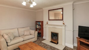 Una sala de estar con un sofá color beige, una mesa de centro de madera, una chimenea, una estantería y un televisor sobre un mueble de madera.