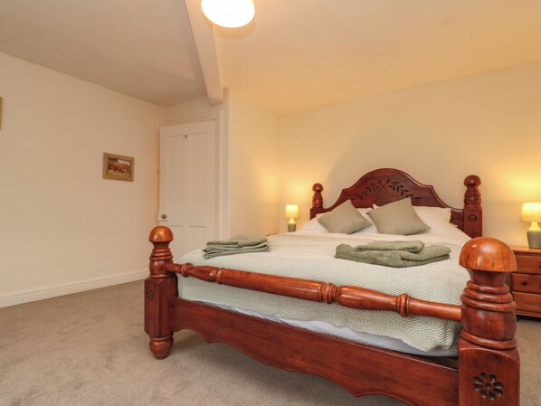 Una cama bien tendida con estructura de madera y cubrecama verde, una mesita de noche con lámpara, y una puerta al fondo.