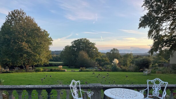 Eine Garten-Terrasse mit weißen schmiedeeisernen Stühlen und einem Tisch, mit Blick auf einen gepflegten Rasen und Bäume.