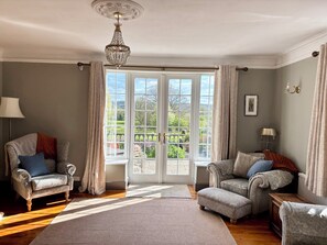 A room with two armchairs, a sofa, a chandelier, and a large window with a view of a garden. - Humphreston House (Bristol)