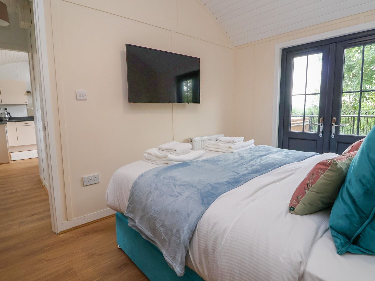 A bedroom with a bed, a TV mounted on the wall, a door leading to a balcony, and a visible kitchen area.