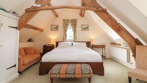 A bedroom with a sloped ceiling, a large bed, a wooden bench, a nightstand, a dresser, and a window with curtains. - Highgrove Barn (Stroud)