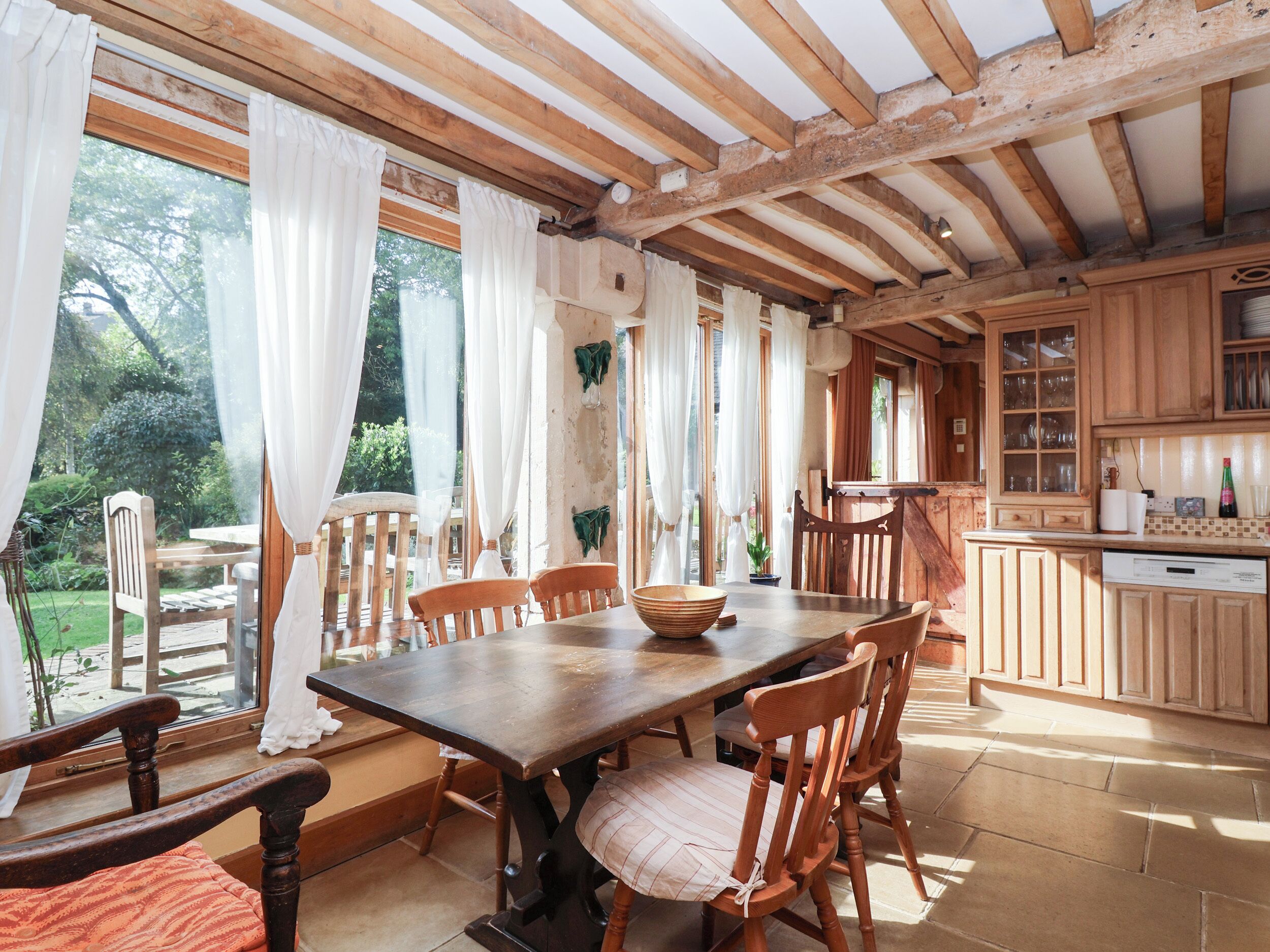 Un comedor rústico con una mesa y sillas de madera, ventanas amplias con cortinas blancas y una cocina con armarios de madera y lavavajillas.