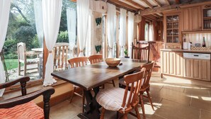 A rustic dining area with a wooden table and chairs, large windows with white curtains, and a kitchen with wooden cabinets and a dishwasher. - Highgrove Barn (Stroud)