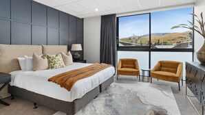 Een moderne hotelkamer met een groot bed, twee fauteuils, een tafeltje en uitzicht op een brug en heuvels.